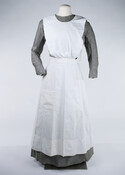 World War I nurse's uniform consisting of a dress and an apron. The gray dress is full-length, collarless and has long sleeves. It fastens at the left shoulder, left front, and waist with sets of four plastic buttons. Worn under a white apron with bib neck. Bib has small pleats at waist and waistband fastens…