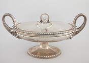 Silver covered tureen standing atop an ovular pedestal. Decorated with a beaded border on base and lip of lid. Two handles are made in the form of serpents with two snake heads and the figured of a bearded man on appearing on either side. This tureen was one of an identical pair that was split…