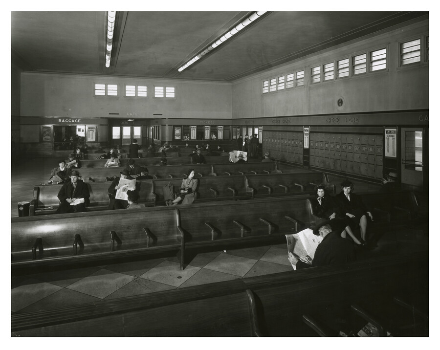 A view of the interior of the Greyhound bus terminal located at 601 North Howard Street in Baltimore, Maryland. Passengers sit waiting for buses, while in the background signage for a baggage area is visible. The terminal was in operation from 1941-1987.