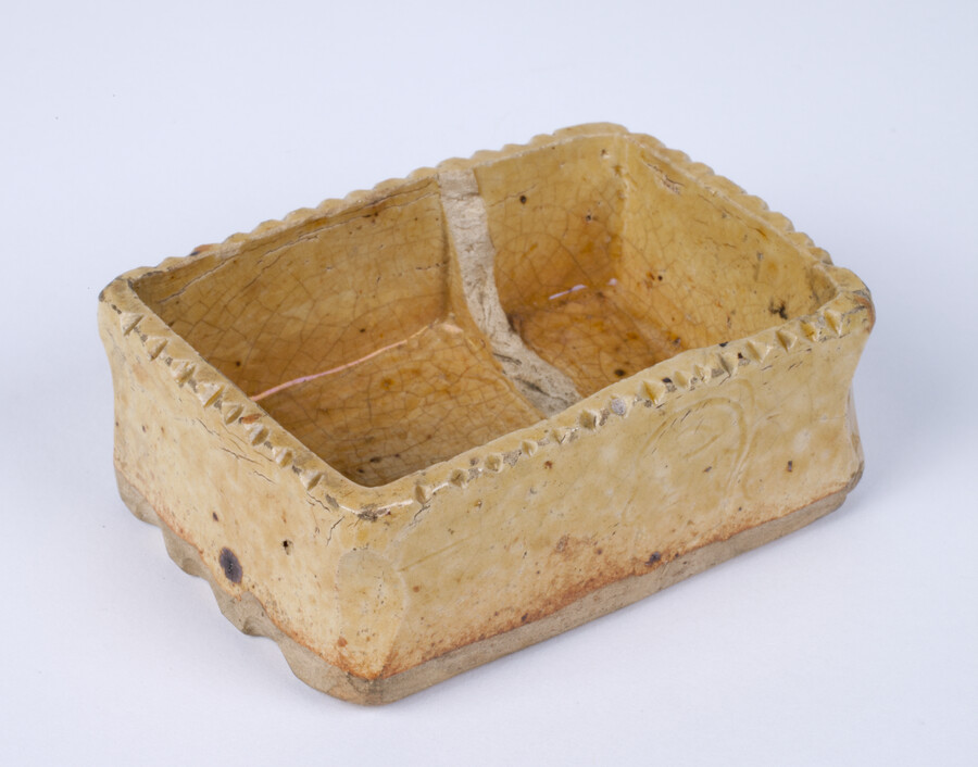 Earthenware rectangular dish with yellow lead glaze embellished with scalloped rim, base and corner edges. Interior divider inside dish now broken. Face incised with date, "1806."