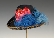 Black felt hat with flat top and wide brim turned up at edges. Edges of brim embellished with looped gold trim. Embellished with blue feathers on both sides and large coral satin bow on one side.