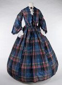 Blue, pink, and green plaid silk taffeta dress. Bodice has scalloped neckline to waist and a hook-and-eye front closure. Collar, inner bodice, and undersleeves are made of white muslin trimmed with eyelet embroidery. Two-tiered pagoda sleeves are edged with blue and black silk fringe and two rows of scalloped taffeta at the top of the…