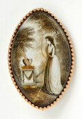 Oval-shaped mourning brooch featuring an image of a woman by a tombstone with heart shapes on its top. Engraved with the initials "M.L." on the verso. The back opens to reveal a sample of hair and the initials "H.M." on glass covering the hair.