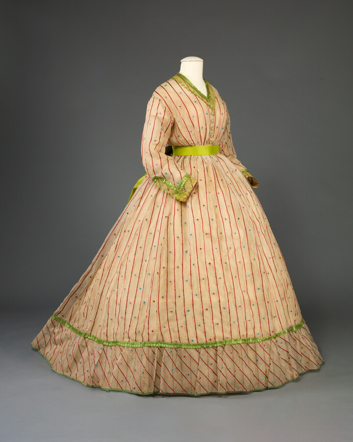 Sheer, gauzy dresses such as this one became popular in the 1860s, especially with younger women. Open weaves, rather than closely-woven silk gowns, were particularly desirable in the summer months. Despite the prettiness of these sheer, light dresses, they lacked the vibrant colors of their silk taffeta contemporaries. To add color, the dresses were trimmed…