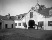 View of two horses and handlers standing outside the stables on the grounds of Brooklandwood (or Brookland Wood), an historic home in the Brooklandville community of Baltimore County, Maryland. Built in the mid-1790s on land purchased by Charles Carroll of Carrollton, Brooklandwood became the country home of Richard and Mary Carroll Caton. The estate was…
