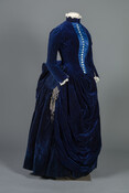 Navy blue velvet two-piece dress. The velvet and silk satin bodice closes in the front with pearl buttons and hooks and eyes. The skirt features decorative bead-work, swagged fabric in the front, and an interior bustle.