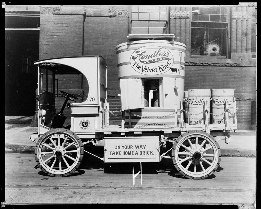 A Hendler's ice cream truck with an oversized wooden ice cream churn. The churn features the Hendler's logo with the company's catch phrase, "The Velvet Kind." Beneath the truck bed is the wording "On Your Way Take Home A Brick." The truck is numbered 70 and is parked in front of the Hendler's building at…