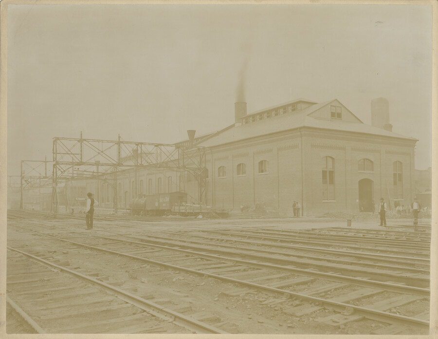 View, looking northeast from the 300 block of West Henrietta Street, of the Baltimore Belt Line's South Howard Street powerhouse located in Baltimore, Maryland. People stand on the nearby train tracks. The building contained 500-Kw electrical generators that produced the power for electric locomotives to pull Baltimore & Ohio Railroad trains through the 1.4-mile long…