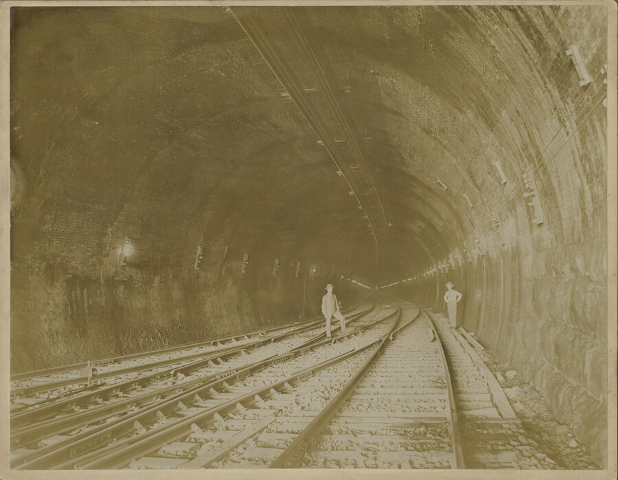 Two people stand inside the Howard Street Tunnel in Baltimore, Maryland. Constructed by the Baltimore Belt Line, which was a wholly-owned subsidiary of the Baltimore & Ohio Railroad, the tunnel was built between 1890 to 1895 and was the longest in the B&O system at 1.4 miles. The Baltimore Belt Line was created in order…