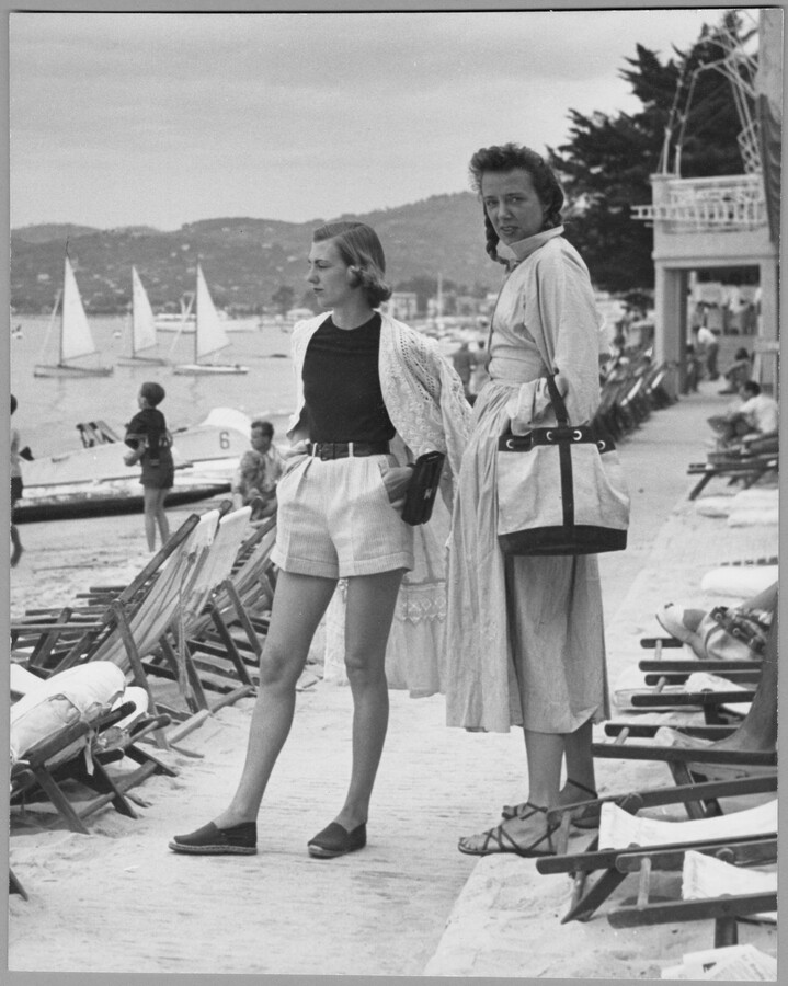 Claire McCardell, accompanied by a younger person, standing amidst beachfront lounge chairs.