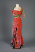 Evening dress constructed from changeable coral and gold silk velvet. It consists of a bodice and a skirt. The bodice features an asymmetrical trim of metallic gold lace, green silk thread, and assorted glass and metal beads. It is from Jeanne Paquin's Winter 1911 collection.