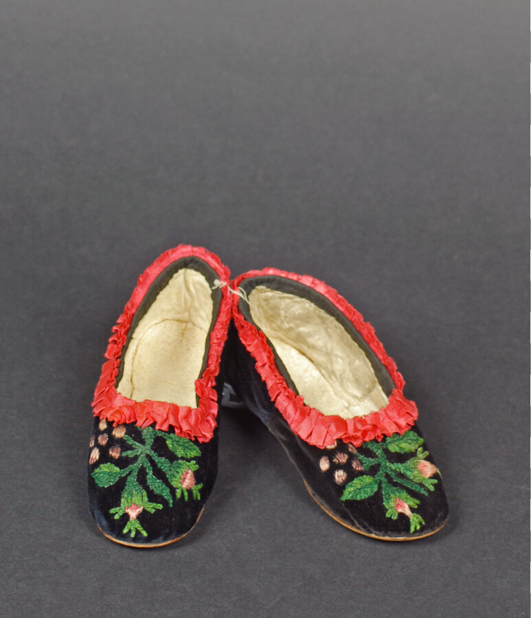 Pair of child's black velvet shoes. Toes and heels of each shoe are embroidered with green chenille and purple and pink silk thread to depict a floral motif. Red ruched silk trim decorates edging of flats. Insoles are white kid leather.