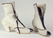 White buckskin leather ladies' boots with low heel made and sold by Hutzler's. Tongue, tip, and seams covered in contrasting black patent leather. Lined with white cotton and kid leather.