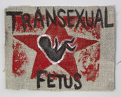 Handmade patch representing the donor's band, Transexual Fetus. Each member of the band was a member of the trans community. The patch features the name of the band with the first word at the top and the second at the bottom, written in black paint. A picture of a fetus in black paint outlined in…