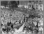 A band on stage in front of a crowd gathered at Mount Vernon Place in Baltimore, Maryland, to celebrate the arrival of the Marshal of France, Joseph Jacques Césaire Joffre, and the French Mission. Joffre visited the city as part of a French delegation touring the country after the United States entered World War I.