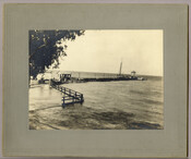 View of the pier on the Chesapeake Bay in Tolchester, Maryland. Figures can be seen walking on the pier and a boat is docked at the end of it. Tolchester was the site of the Tolchester Beach Amusement Park, which consisted of a bathing beach, amusement park, racetrack, and hotel, and was in operation from…