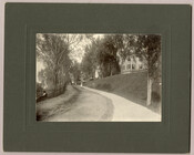 A road winding next to a hill with houses in Tolchester, Maryland. A figure sits next to the road. Tolchester was the site of the Tolchester Beach Amuseument Park, which consisted of a bathing beach, amusement park, racetrack, and hotel, and was in operation from 1877 to 1962. The Kent County park was a popular…