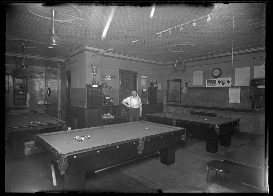 Interior view of a billiards room with unidentified man standing between the tables.