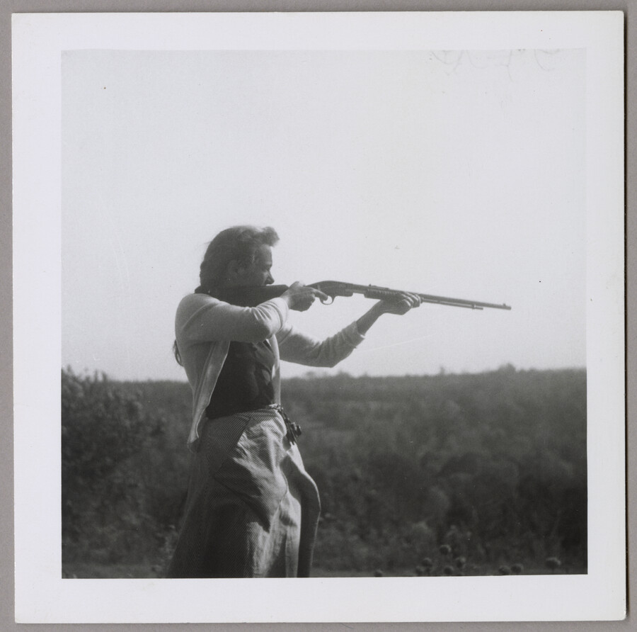 Claire McCardell stands in a field holding what appears to be a shotgun aimed out of the camera's frame. McCardell was a renowned fashion designer from Frederick, Maryland.
