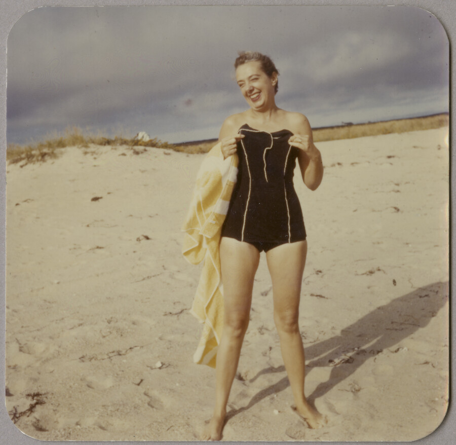 Claire McCardell standing on a beach wearing a black bathing suit with white piping. She smiles at the camera and holds a towel. Claire McCardell was a renowned fashion designer from Frederick, Maryland.