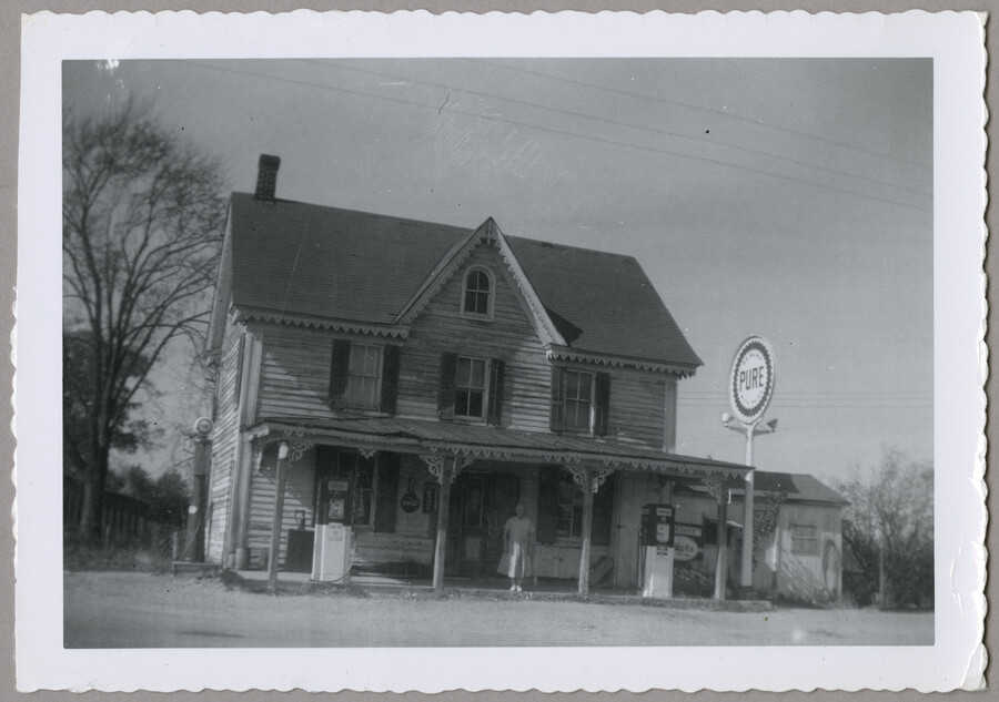Exterior view of the general store run by the Lesage family, located at the corner of Route 298 and Porters Grove-Melitota Road in Melitota, Maryland. A woman stands on the porch of the building.