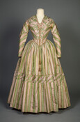 Fashions of the 1840s characteristically favored more subtle and subdued silhouettes, yet the meticulous manipulation of fabrics and patterns maximized a dress’s visual interest. This pale green and lilac striped dress showcases the masterful sewing skills used to manipulate the fabric. Clever pleating at the bodice creates a chevron design and the three strips of…
