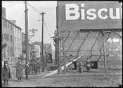 View looking east at a group of children on the southeast corner of E. Pratt Street and S. Exeter Street in Baltimore, Maryland. Part of the word "Biscuit" can be seen painted on the side of the building.