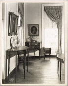 View of a room in the Homewood estate, including a variety of tables and decor, and two framed portraits. Homewood was built between 1801 and 1806 as a country home for Charles Carroll, Jr., son of Charles Carroll of Carrollton who was a signer of the Declaration of Independence. The Federal-period Palladian home was in…
