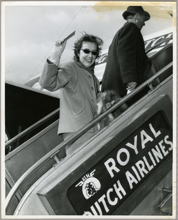 Photograph of American fashion designer Claire McCardell boarding a Royal Dutch Airlines airplane with an unidentified man and waving back at the camera.