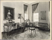 View of a sitting room in the Homewood estate, with a bench, chairs, mirror, framed portraits, rug, harp, and piano. Homewood was built between 1801 and 1806 as a country home for Charles Carroll, Jr, son of Charles Carroll of Carrollton who was a signer of the Declaration of Independence. The Federal-period Palladian home was…
