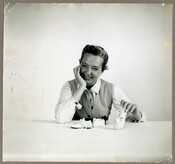 A portrait of American fashion designer Claire McCardell seated at a table with baby shoes, smiling with her head on her hand.