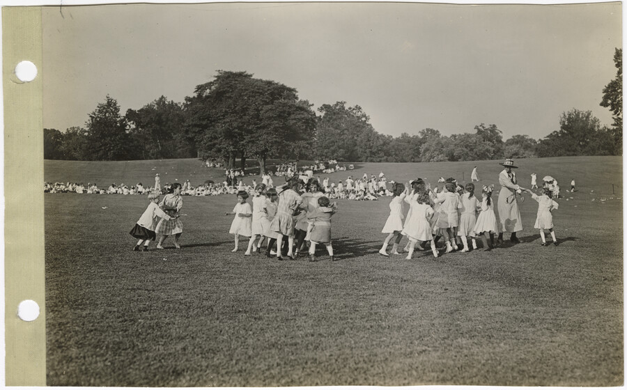 Children play a game of 'King Gaul' on the lawn of Druid Hill Park, Baltimore, Maryland; while an adult supervises. Large groups of children and some adults are in the background. In the far distance trees, and possibly a wooded area line the edge of the field.