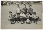 The children's champion volleyball team is posed, seated, with a ball placed on top of a hat, in front of the group. They are positioned on a volleyball field, with spectators in the background. The group of eight are likely all boys.