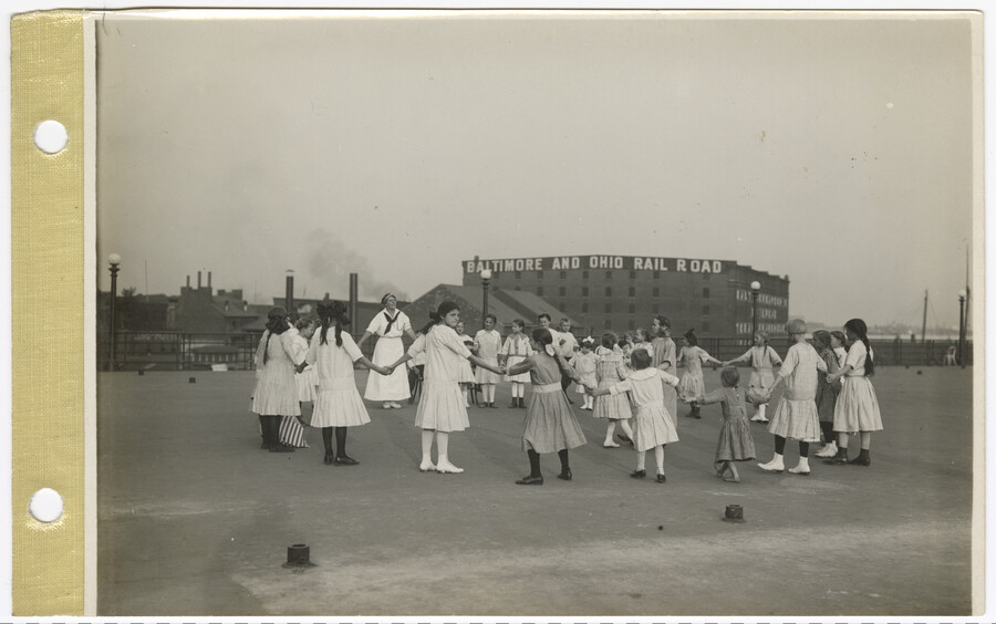 A view of children positioned in a circle, possibly dancing the 'Herr Schmidt', a traditional German dance. One adult is present. A sign for 'Baltimore and Ohio Rail Road' appears on a warehouse behind the park. The waterfront is on the right and industrial buildings are on the left in the background of the image.