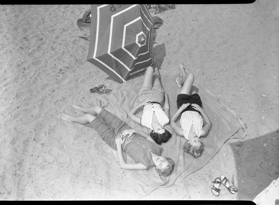 An overhead view of three beach-goers lying on a blanket at Sandy Point State Park in Annapolis, Maryland. A striped umbrella is set in the sand nearby. Opened in 1952, the park is a public recreation area on the Chesapeake Bay and is known for the popularity of its swimming beach.