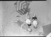 An overhead view of three beach-goers lying on a blanket at Sandy Point State Park in Annapolis, Maryland. A striped umbrella is set in the sand nearby. Opened in 1952, the park is a public recreation area on the Chesapeake Bay and is known for the popularity of its swimming beach.