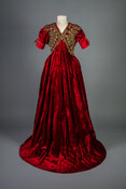 Red velvet evening coat embellished with bronze and olive green silk satin embroidery and cording. It features a plunging neckline, short puffed sleeves, and an open front.