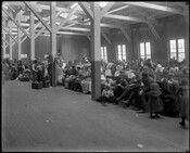 A waiting area for immigrants at the immigration pier in the Locust Point area of Baltimore, Maryland. Immigrants were required to wait here, as they were not allowed to leave the pier unless directly boarding a designated train. A sign on the wall behind them has "No smoking" written in three languages: English, German, and…