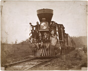 Portrait of a group of men and women standing and sitting on the cowcatcher at the front of a train, flanked by two American flags. The individuals in the photo are presumed to be from a larger group of artists and businessmen invited to make an excursion by Baltimore and Ohio Railroad train starting from…