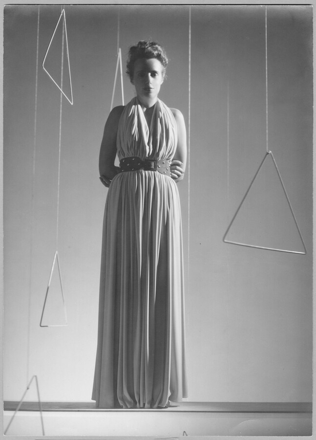 A full-body portrait of fashion designer Claire McCardell standing among metal triangles suspended from the ceiling. McCardell's attire is reminiscent of her “Monastic” dress, a style defined by its loose shape and lack of a defined waistline until the wearer used a belt to cinch in the fabric.