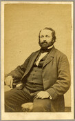An undated portrait of an unidentified man. The man is seated with his right arm resting on a table; he sports a beard and wears a suit.
