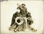 Group portrait of bandleader James Reese Europe and two other musician-soldiers from the military band of the 369th Infantry Regiment commonly referred to as the "Harlem Hellfighters." The three band members pose with a variety of brass instruments in their military uniforms.