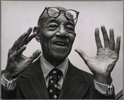 Portrait of American pianist, lyricist, and composer Eubie Blake, possibly taken by Ian Lowrie. Blake holds his hands in the air with his glasses pushed up onto his forehead.