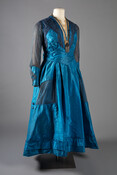 Aniline blue silk taffeta dress with chiffon sleeves and faux pockets, cream net collar and metallic bodice under chiffon with floral beaded embroidery. Connected necklace with large metallic tassel. Made by Stuart & Jackson in Baltimore, Maryland.