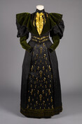 Black two piece dinner dress with high neck and gigot sleeves. Embellished with exaggerated green velvet collar, neon ruffle front, jet beading, and floral design on skirt. French origin, imported by E. Craig. Dated to 1895, Elizabeth Vickers Anderson’s dress presents the cyclical nature of fashion with its voluminous leg-of-mutton sleeves, a style previously popular…