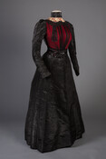A ladies two-piece dinner or reception dress with a fully boned bodice of red silk satin covered in black net gathered at the neck and waist and trimmed with black jet and sequins. The dress features a high collar with cream floral crochet and wide black satin ribbon. Arms are full-length black moire trimmed with…