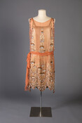 In 1922, the tomb of the Egyptian pharaoh Tutankhamun was discovered, inspiring a worldwide interest in ancient cultures. Egyptian-inspired beadwork, motifs, and colors quickly started appearing in jewelry and clothing, like the scalloped tiers of geometric beadwork in this dress and the gold lamé that shines through the ethereal peach chiffon.