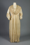 Woman's summer day or tea dress of cream/beige silk gauze. It has a fold over net and lace bib collar, long sleeves with lace and embroidered applique on the sleeves. Bodice has two vertical panels of matching net/lace with embroidery an applique. Gown has darker silk skirt of flowing pleats, bell-shaped scalloped hem with net…