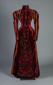 Burgundy velvet tea gown with stripes of gold silk brocade. Floral beaded embroidery on front of skirt. Dress has high neck and full-length sleeves.