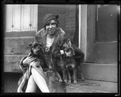 Woman in a fur coat seated on a stoop posing with three small dogs.